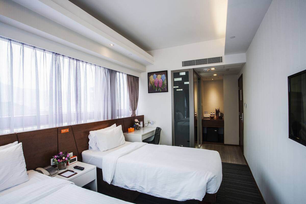A hotel room with two beds, a desk, a phone, a vase with flowers, and a painting on the wall.