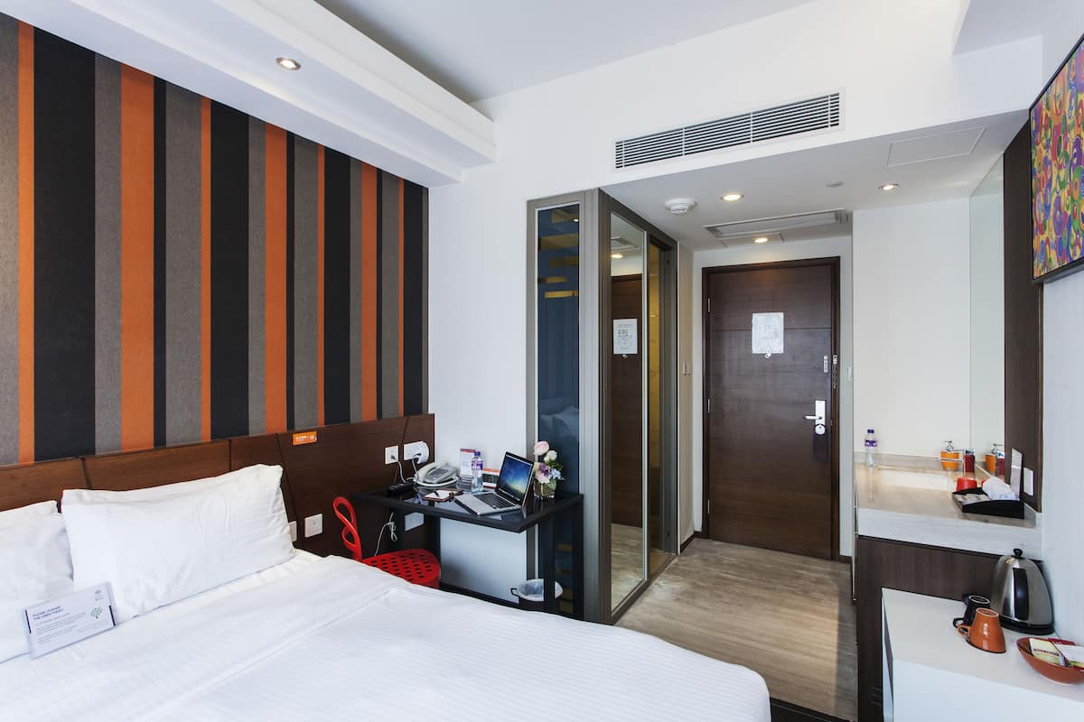 A hotel room with a bed, a desk with a laptop, a red trash bin, and a shower area.