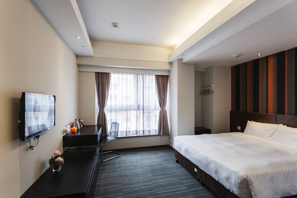 A hotel room with a bed, a desk with a chair, a TV, and a window with curtains.