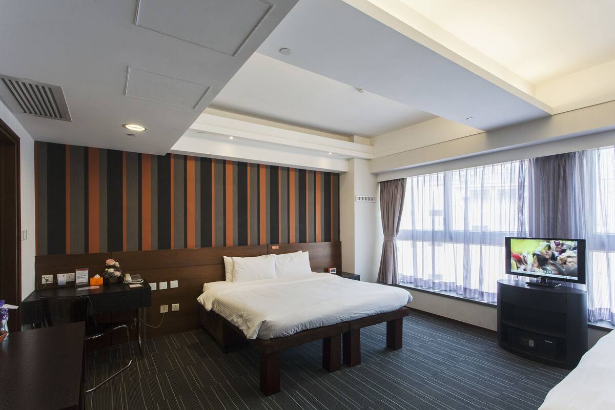 A hotel room with a large bed, a desk, a television, and a window with curtains.