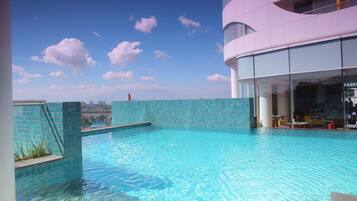 Una piscina en la azotea con agua azul cristalina, rodeada por un edificio moderno con grandes ventanales y un muro de azulejos.