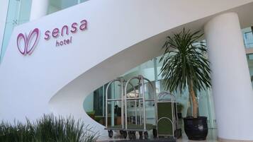 The entrance of the Sensa Hotel with a modern design, featuring a large glass facade and a palm tree in the foreground.