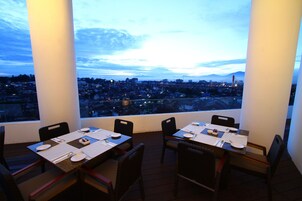 Comedor con mesas dispuestas para dos, con vistas a un paisaje urbano al anochecer.