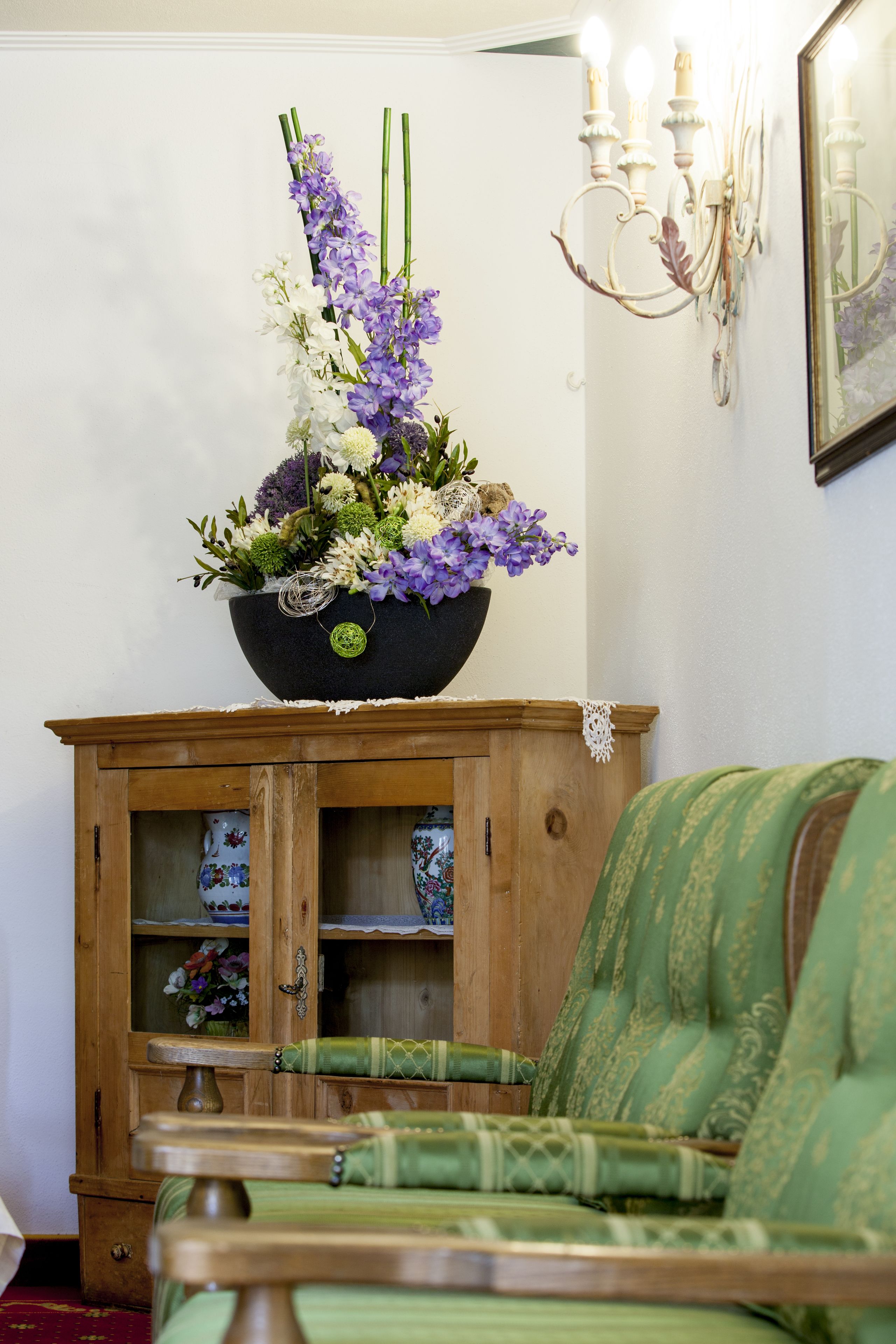 A wooden cabinet with glass doors, a green upholstered chair, and a floral arrangement on top.