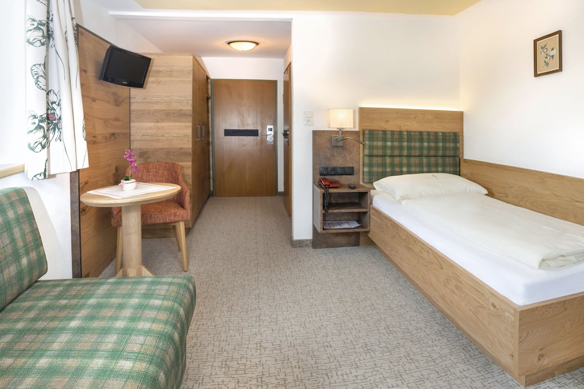 A compact hotel room with a single bed, a small table with a chair, a television mounted on the wall, and a wooden wardrobe.