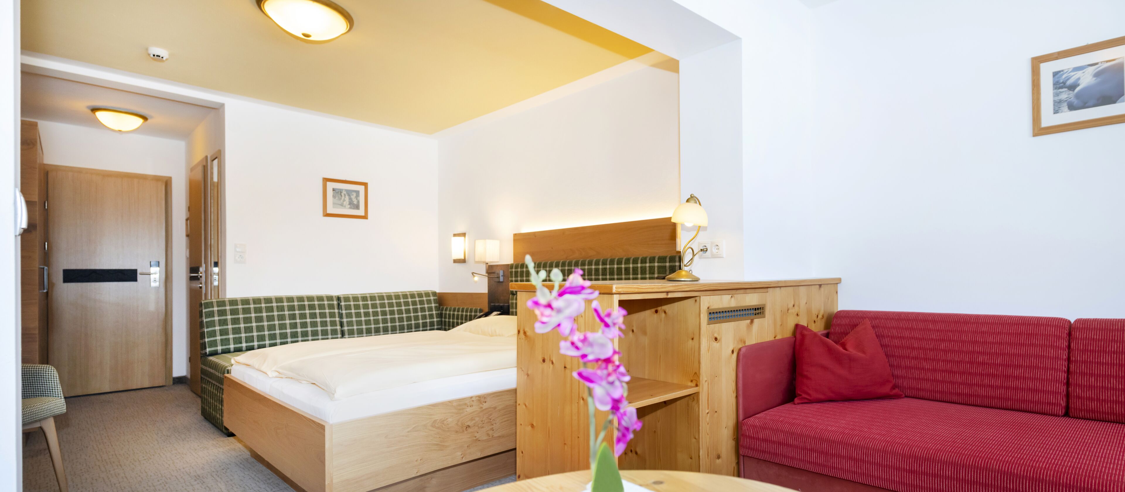 A hotel room with a bed, a red sofa, a desk with a lamp, and a small table with a vase of flowers.