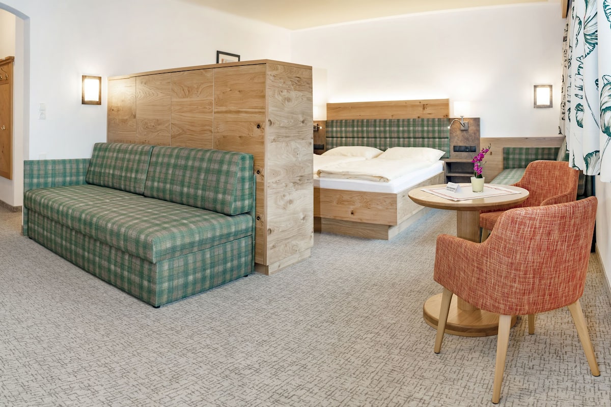 A hotel room with a green plaid sofa, a wooden cabinet, a bed with a green and white checkered headboard, a round wooden table, and two red plaid chairs.