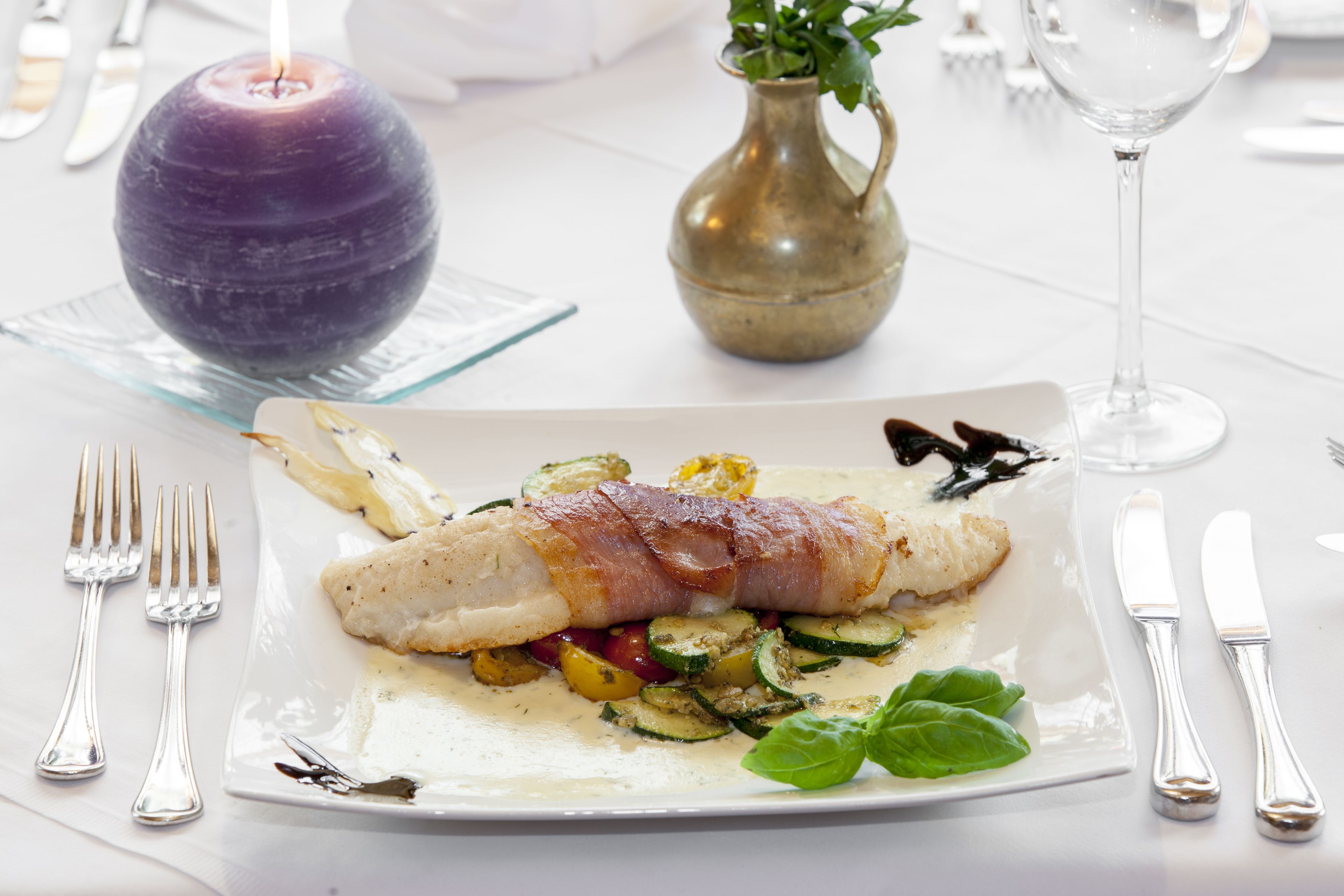 A plate of food with a fish fillet wrapped in bacon, accompanied by vegetables and a basil leaf.