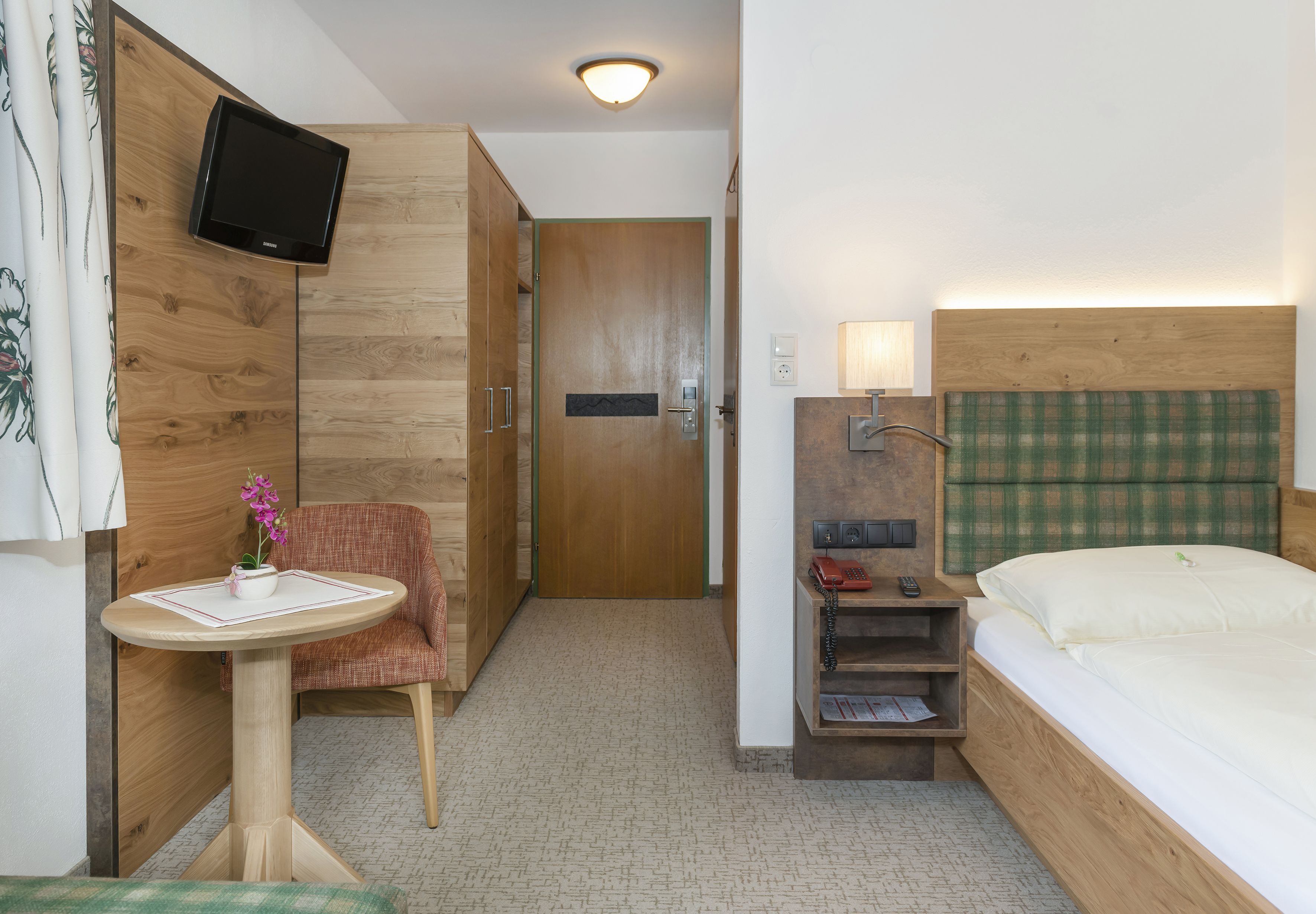 Une chambre d’hôtel avec un lit, une petite table sur laquelle est posé un vase de fleurs, une chaise, un téléviseur fixé au mur et une lampe de chevet.