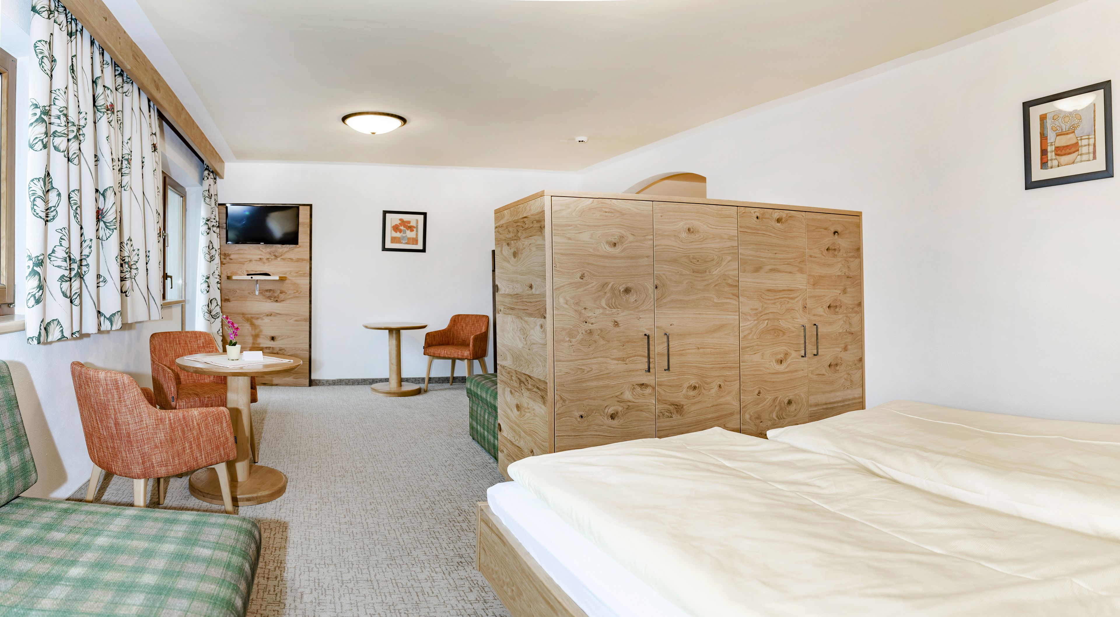 A hotel room with a bed, a wooden wardrobe, a small round table with a chair, a TV mounted on the wall, and a green checkered bench.