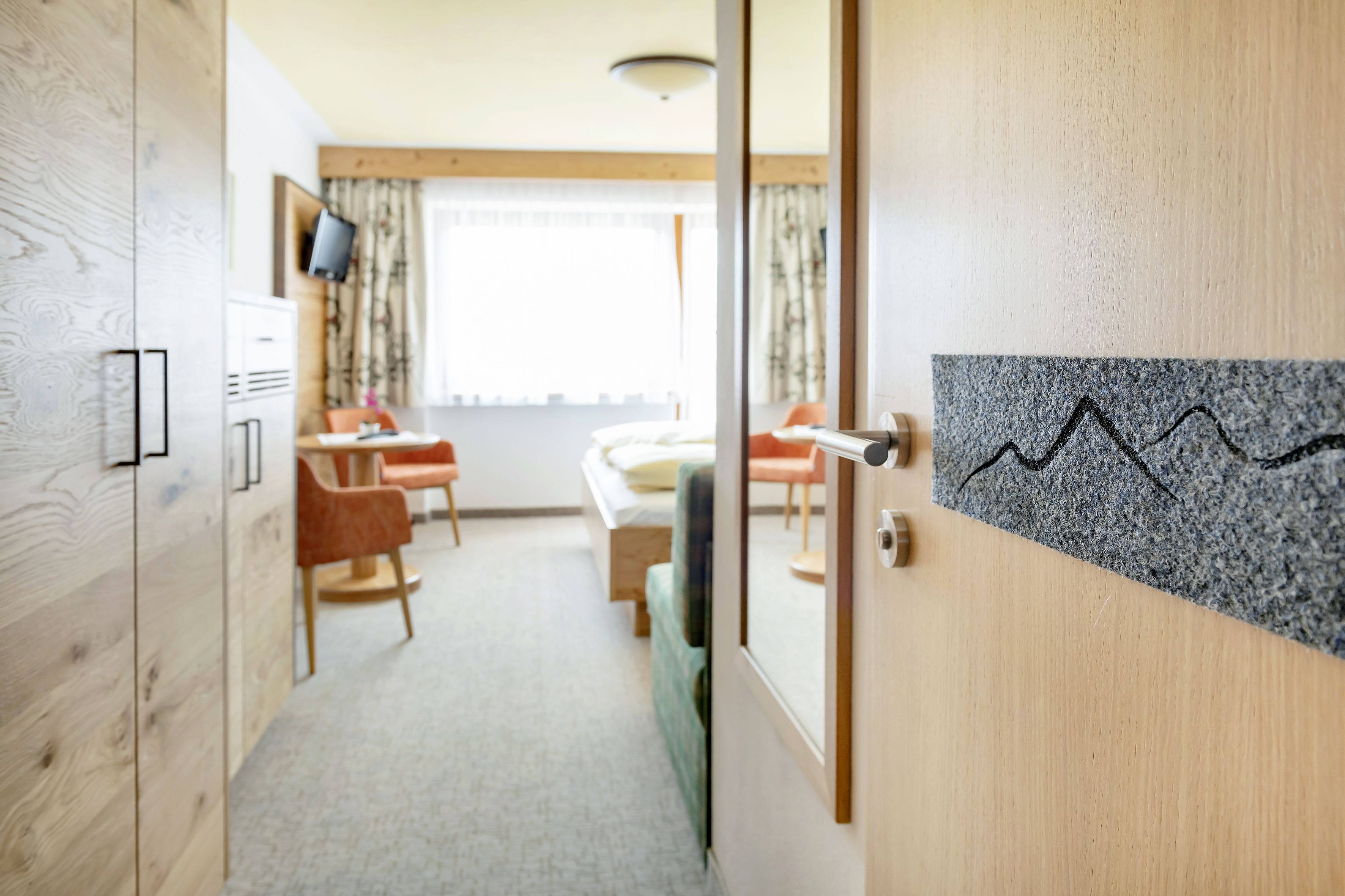 A hotel room with a wooden wardrobe, a bed with a green headboard, a desk with a chair, and a window with curtains.
