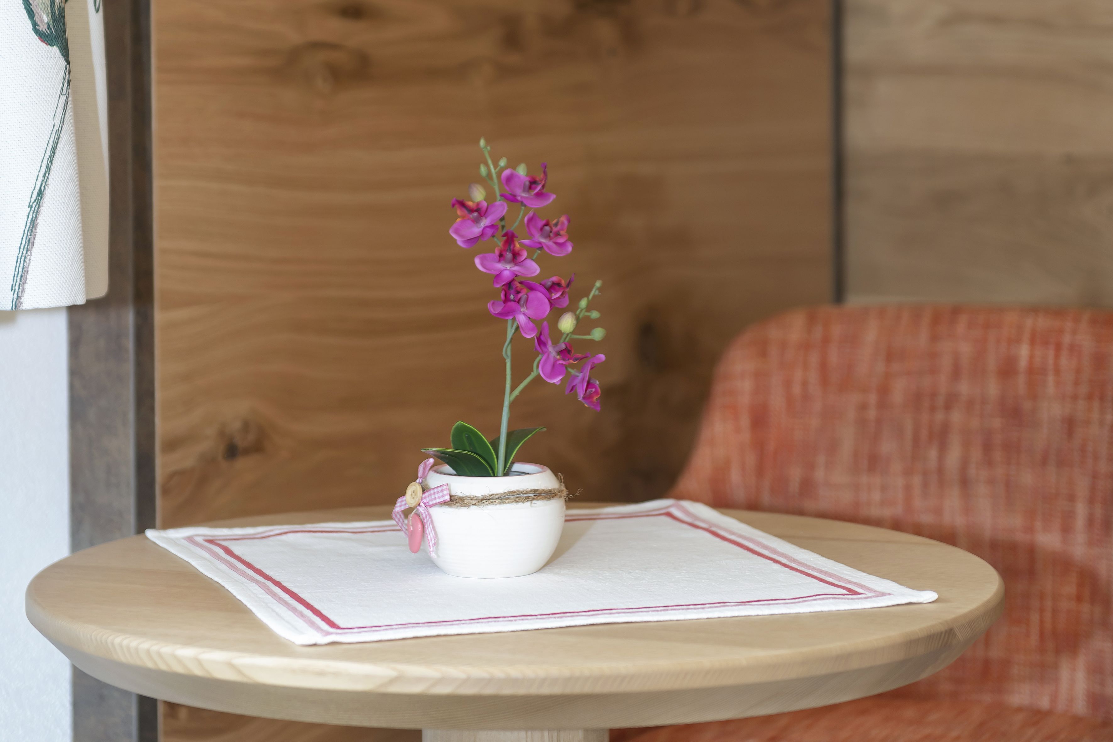 Um pequeno bule branco com um laço rosa sobre uma mesa redonda de madeira com um jogo americano branco.