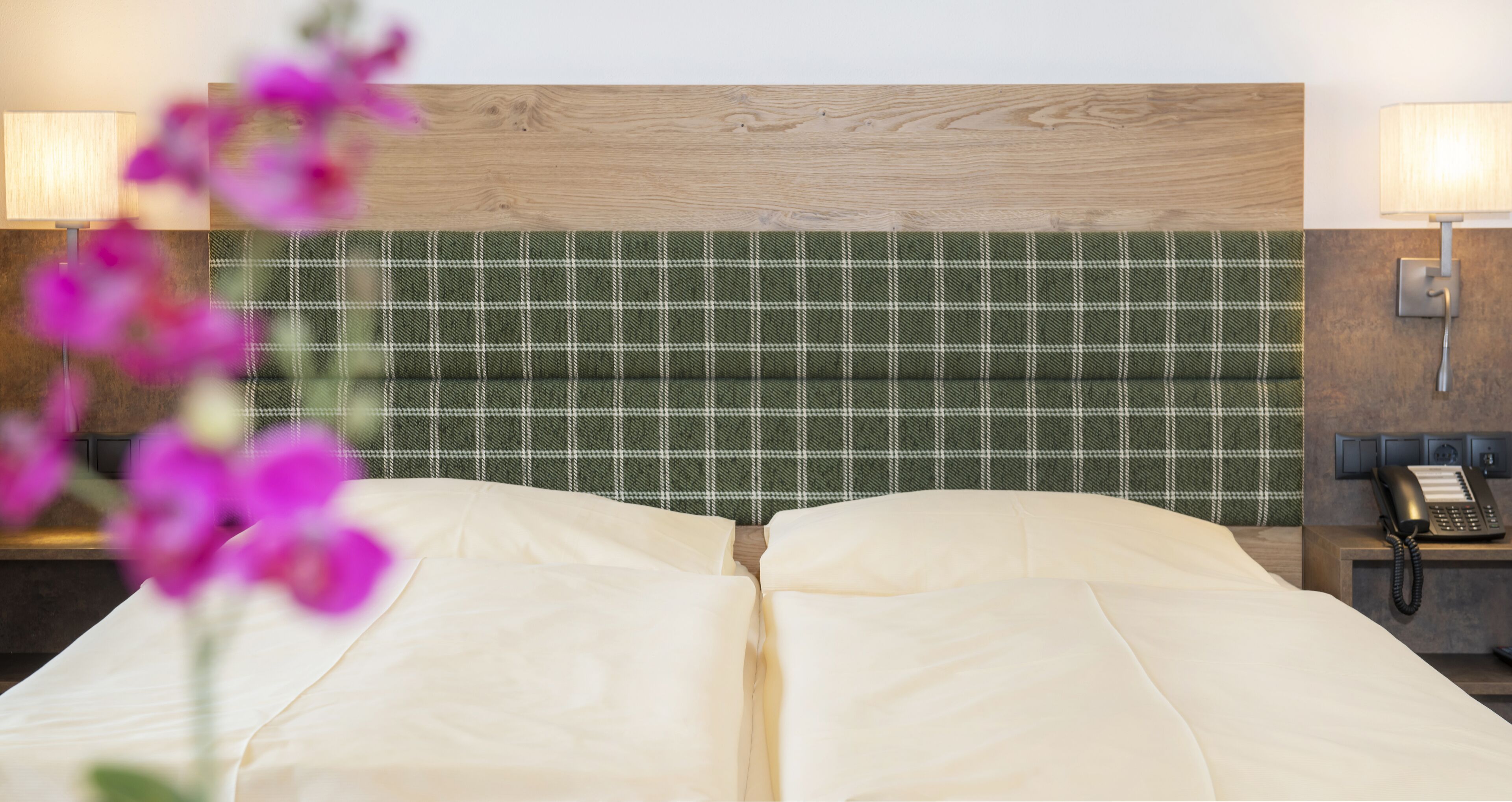 A hotel room with a plaid headboard, two bedside lamps, a telephone, and a bed with white linens.