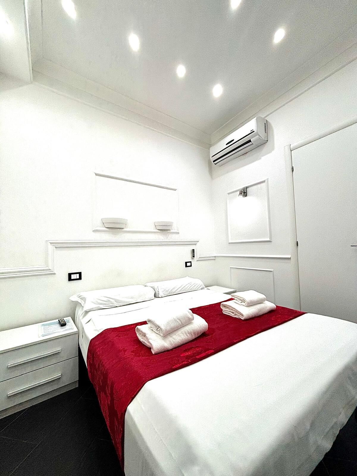 A modern hotel room with a neatly made bed, a bedside table with a lamp, and an air conditioning unit on the wall.
