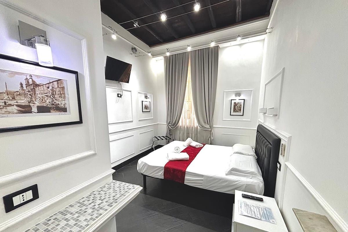 A modern hotel room with a neatly made bed, a flat-screen TV mounted on the wall, a small bedside table with a remote control, and a framed picture on the wall.