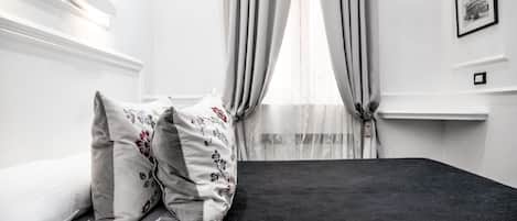 Una cama bien tendida con almohadas blancas y estampadas, un colchón oscuro y una ventana con cortinas de gasa.