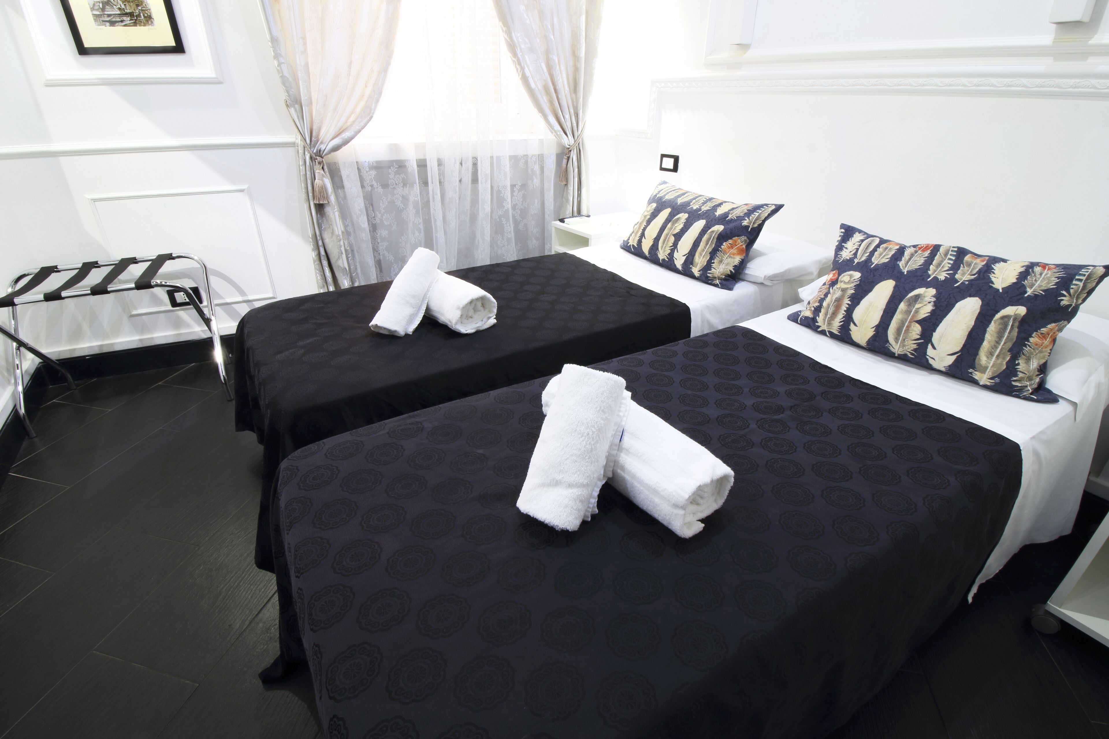 A hotel room with two beds, each with a black patterned bedspread and white towels. There is a small white table and a chair, a window with sheer curtains, and a framed picture on the wall.