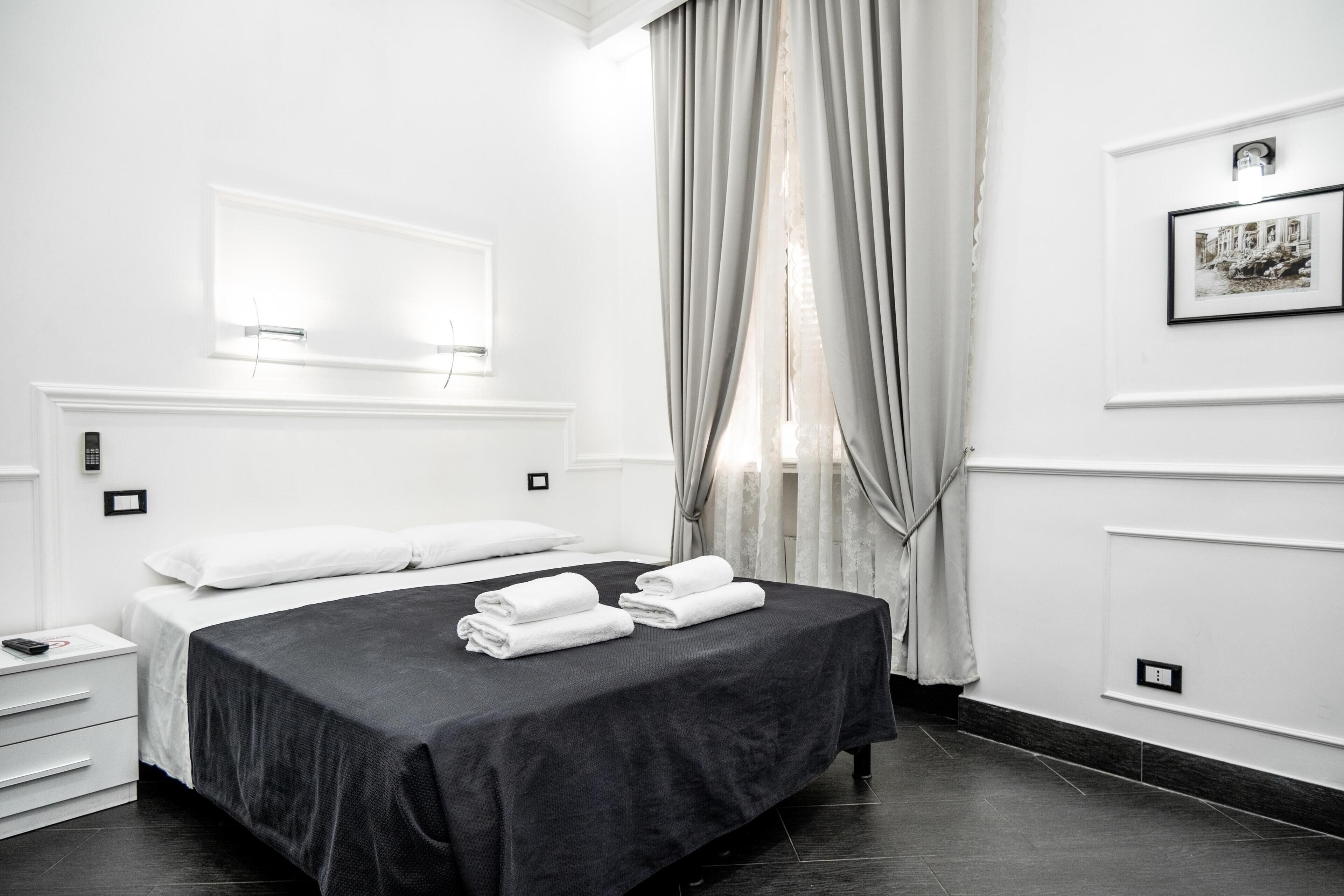 A modern hotel room with a large bed, a bedside table, a nightstand, a wall-mounted light, a framed picture, and a window with curtains.