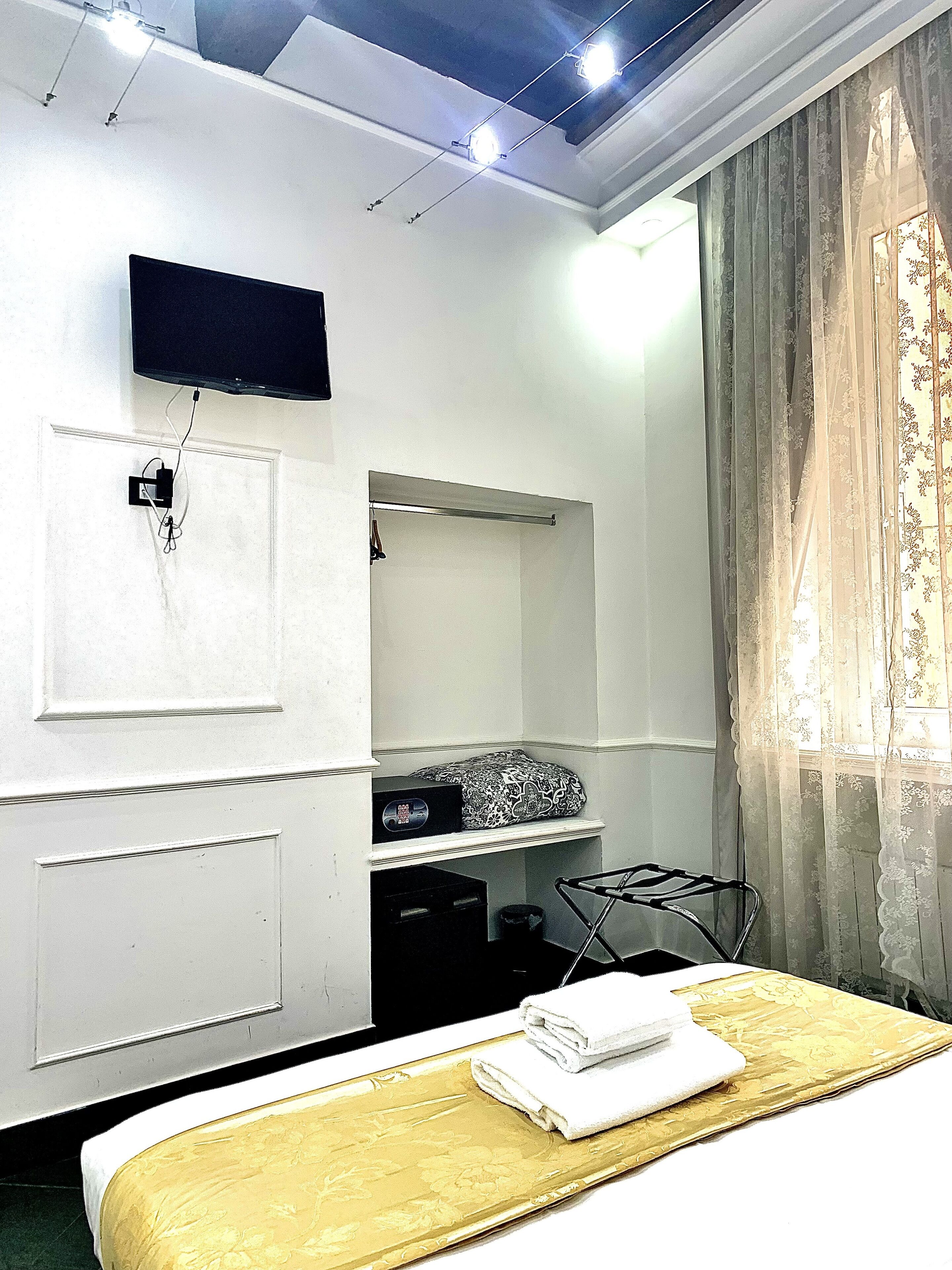A hotel room with a bed, a flat-screen TV mounted on the wall, a shelf with a patterned pillow, and a window with sheer curtains.