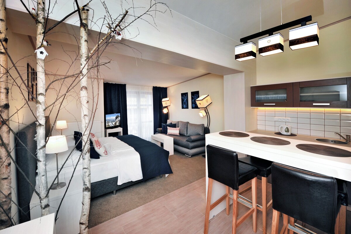 Habitación de hotel con cama, sofá, zona de comedor con mostrador de cocina y ventana grande.