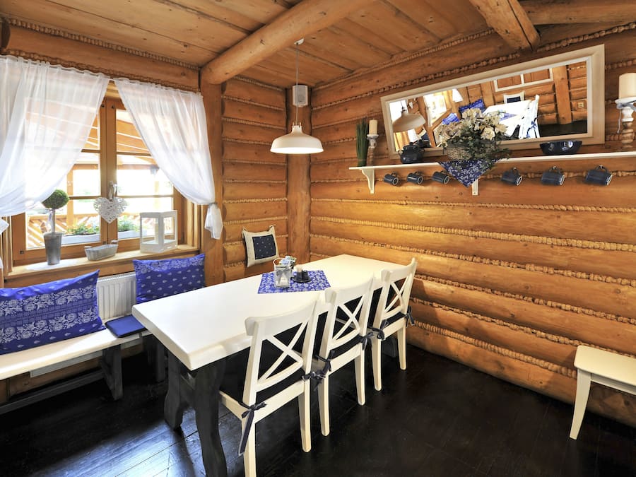 A wooden cabin interior with a dining table, four chairs, a bench with blue cushions, a hanging light fixture, and a mirror on the wall.