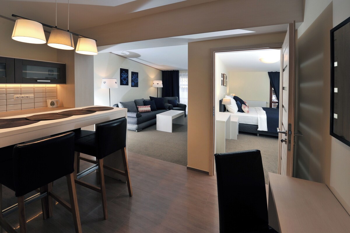 Una habitación de hotel moderna con cocina americana, sala de estar con sofá y mesa de centro, y un dormitorio independiente con cama y mesita de noche.