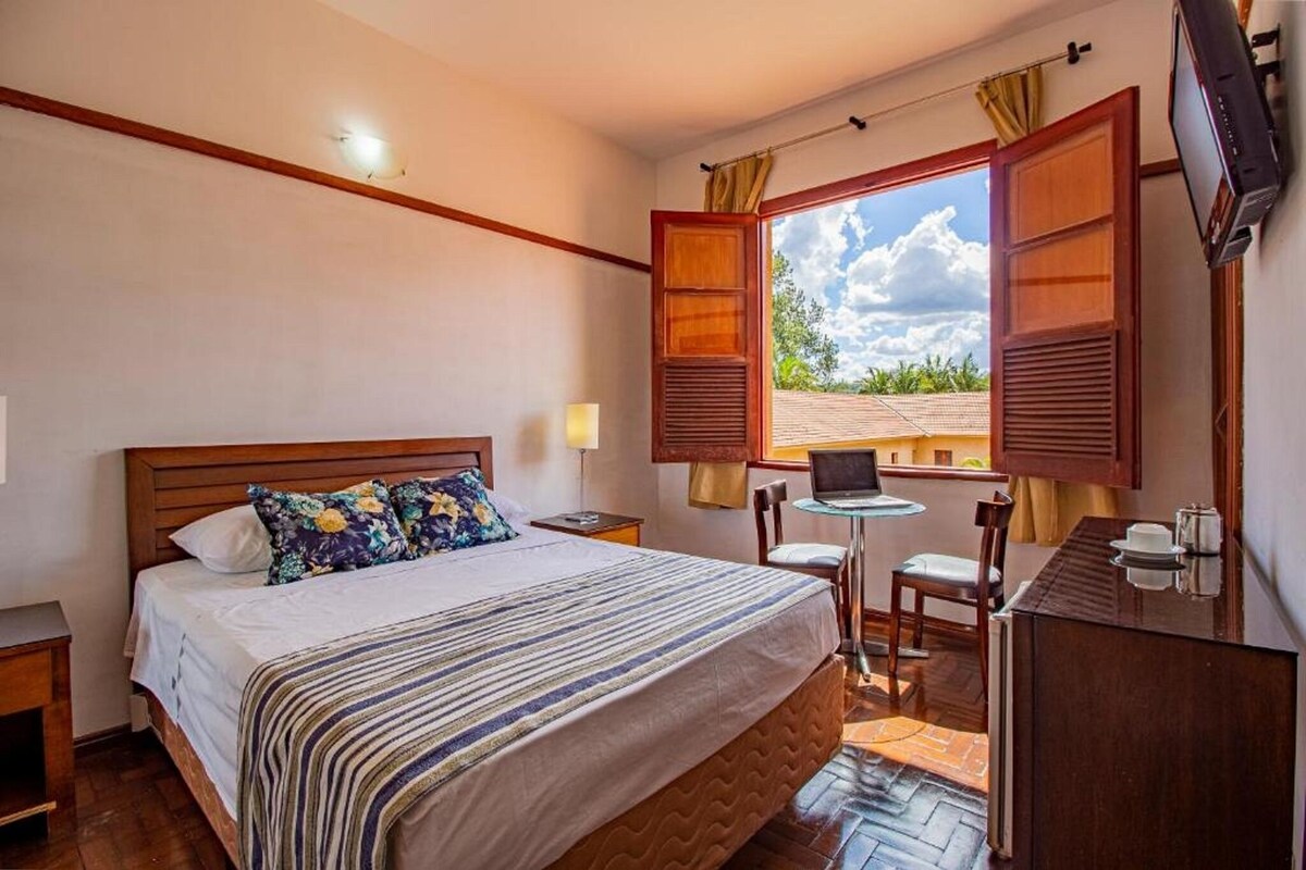 A hotel room with a bed, a desk, a chair, a TV, and a window with open shutters.