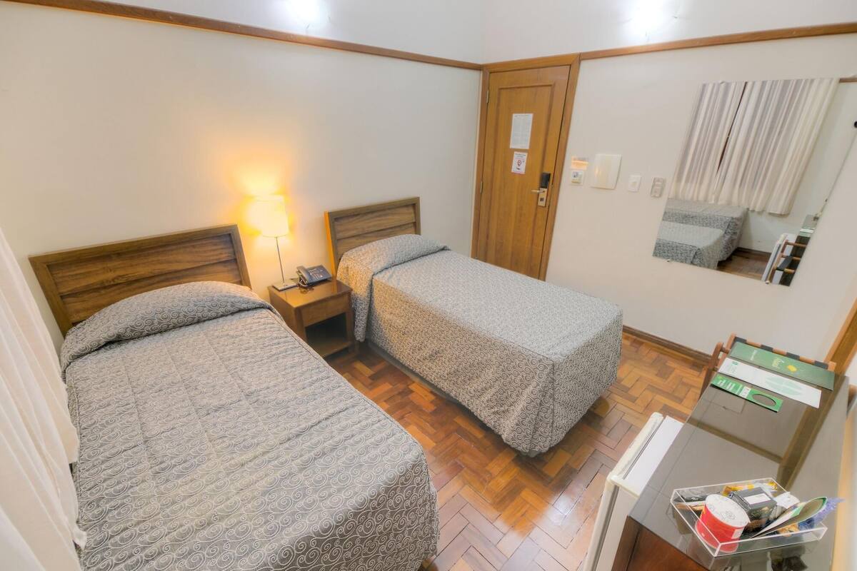 A room with two beds, wooden headboards, a nightstand with a lamp, a mirror, and a small desk with a mirror and some items on it.