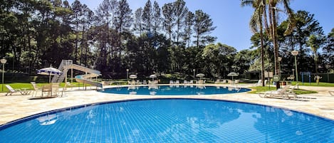 Relax by the serene pool surrounded by lush greenery and tall trees, featuring a rectangular pool with lounge chairs and umbrellas.