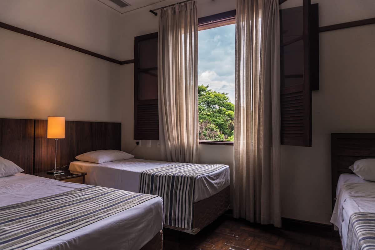 A hotel room with two single beds, a nightstand with a lamp, and a window with curtains.