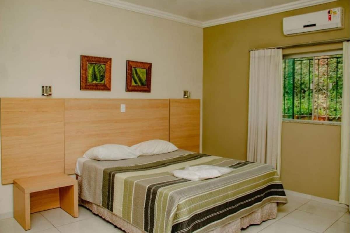 A bedroom with a wooden headboard, two framed pictures on the wall, a window with a view of greenery, and a small wooden stool.
