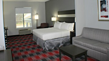 Habitación de hotel con cama, sofá, mesa de centro, escritorio y silla. Cuenta con ventana con persianas, lámpara y un cuadro en la pared.