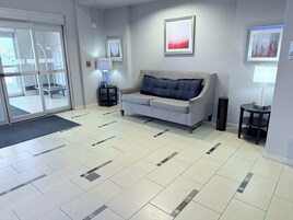 Un hall de hotel con un sofá gris, una mesita con una lámpara y obras de arte enmarcadas en la pared.
