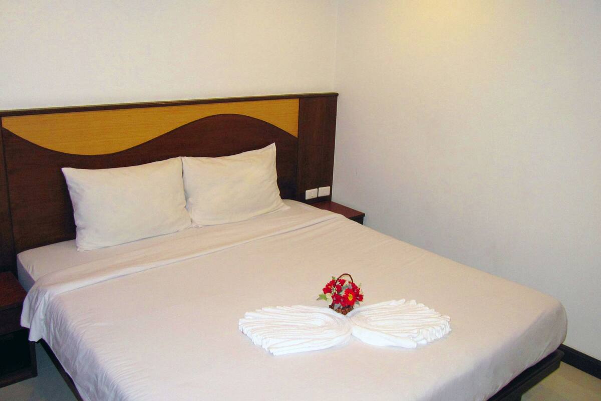 Ein ordentlich bezogenes Bett mit weißen Leinen, einem hölzernen Kopfteil und einer kleinen roten Blumenkomposition darauf.