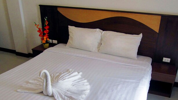 A neatly made bed with a swan-shaped towel in the center, a wooden headboard, and a nightstand with a vase of flowers. - Nina's Guest House 2 (Patong)