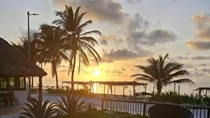 Una piscina con muelle de madera, rodeada de palmeras y un edificio con techo de paja, con vistas a una masa de agua al atardecer.