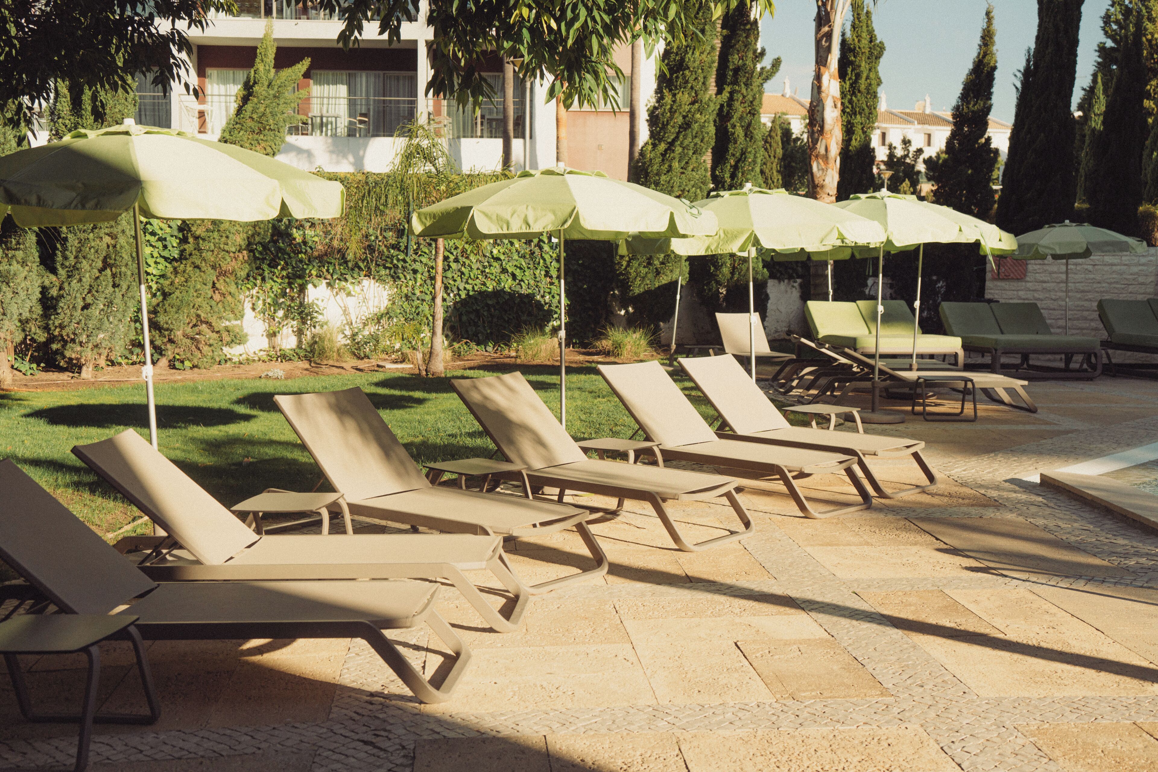 A poolside area with lounge chairs and umbrellas, surrounded by greenery and a building in the background.