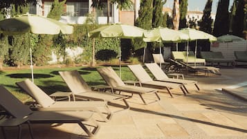 A poolside area with lounge chairs and umbrellas, surrounded by greenery and a building in the background.