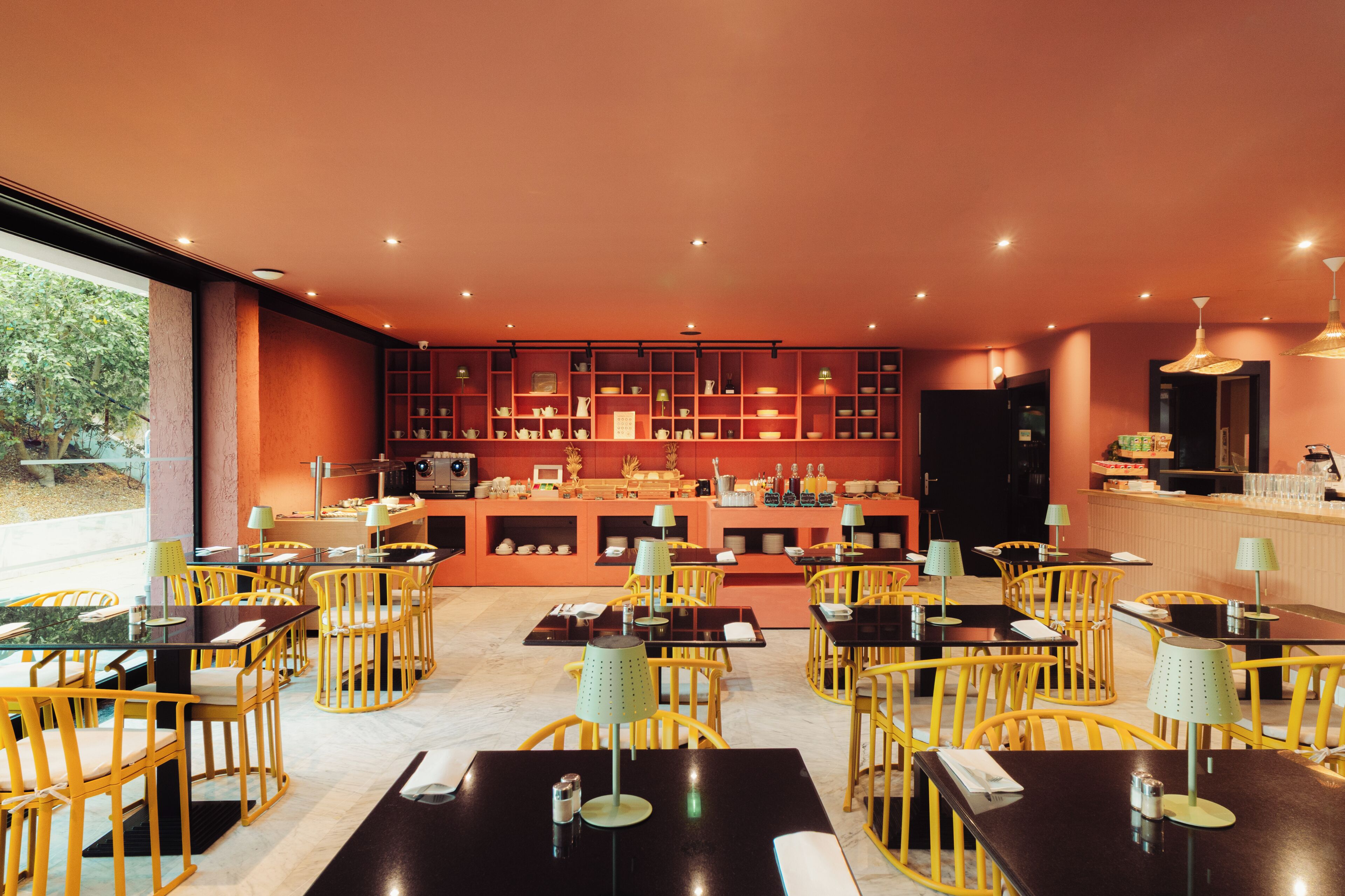A restaurant with yellow chairs and black tables, a bar area, and a view of greenery outside.