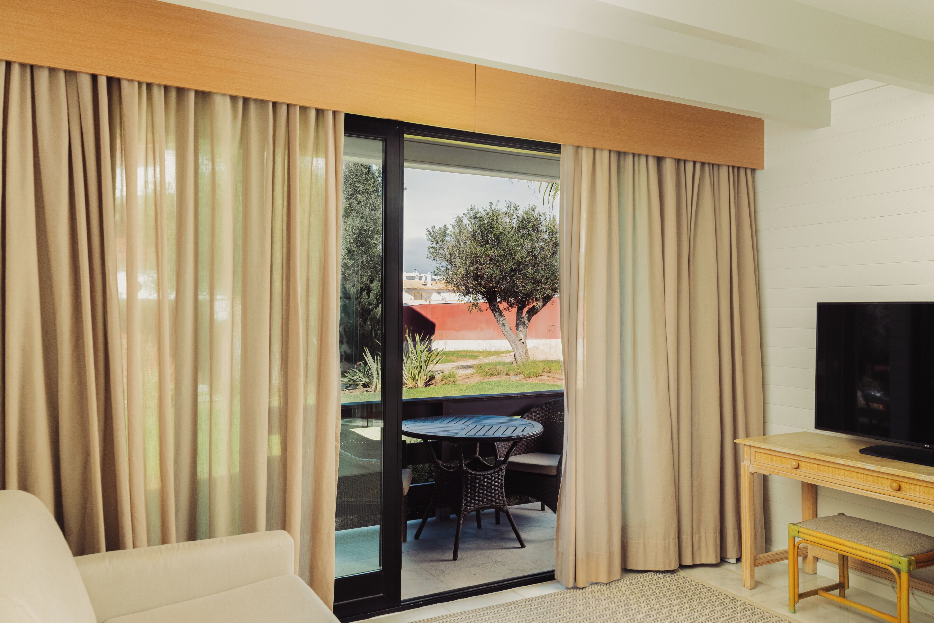 Una habitación con sofá, televisor sobre un mueble de madera, una mesa con sillas y un ventanal corredero que da a un espacio exterior con un árbol y un edificio.
