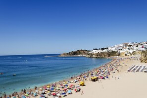 A beach with numerous umbrellas and people, a clear blue sky, and buildings on a hillside in the background.