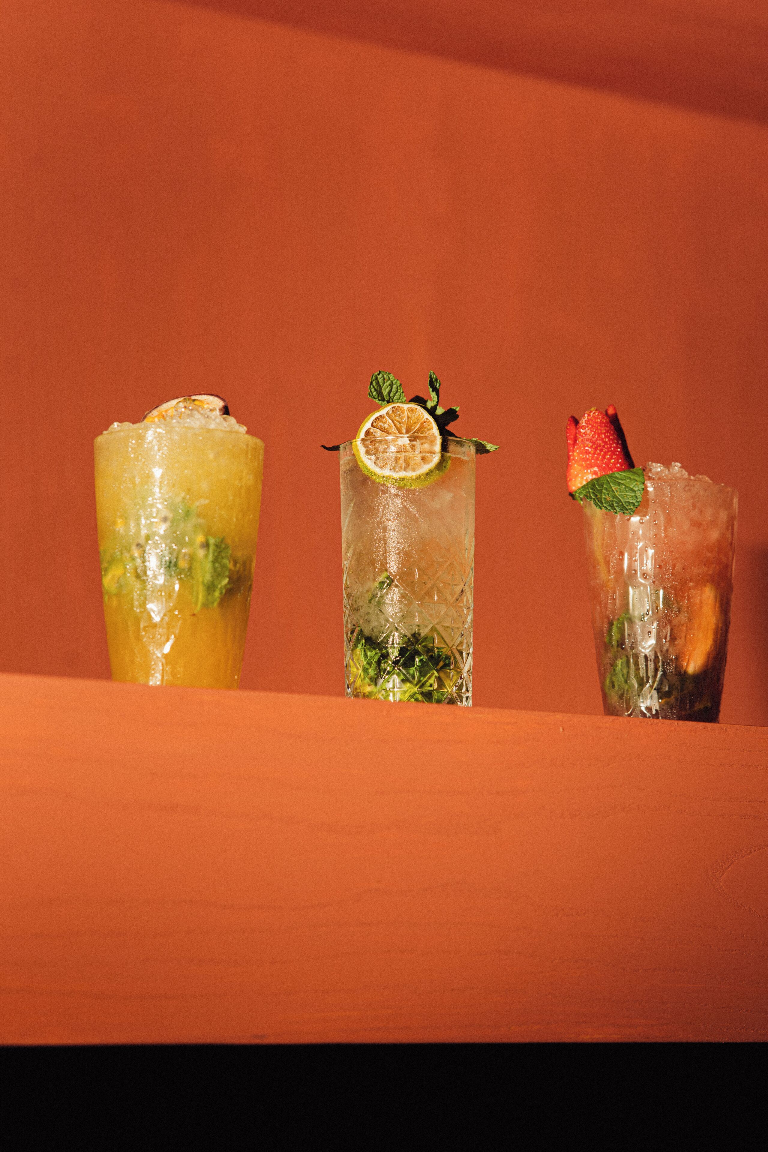 Three glasses of iced drinks with different garnishes on a wooden surface against an orange background.