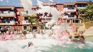 Et poolområde med flere personer, der nyder vandet, omgivet af bygninger med altaner og en klar himmel.
