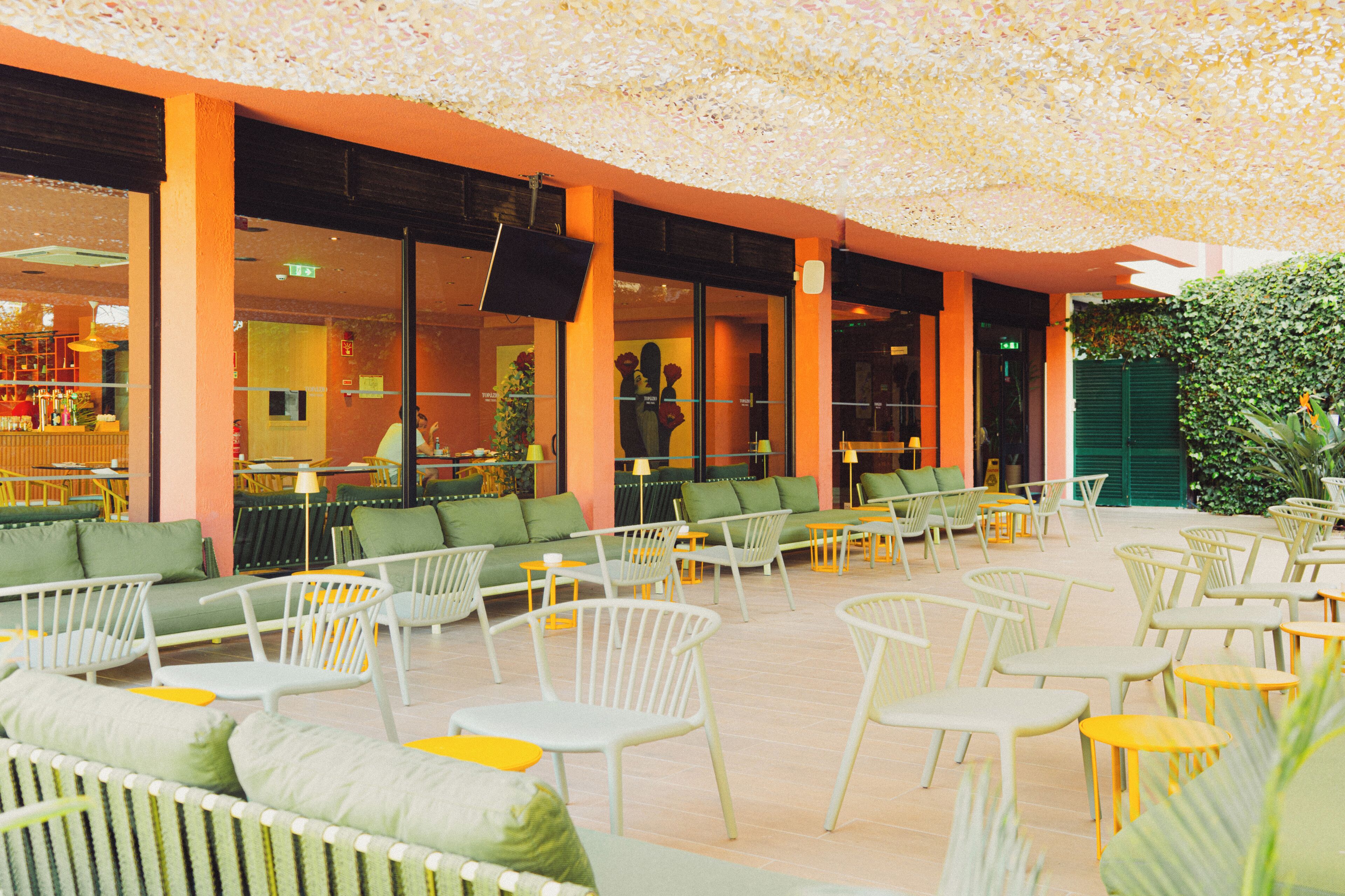 An outdoor seating area with chairs and tables, a bar in the background, and a green wall with plants.