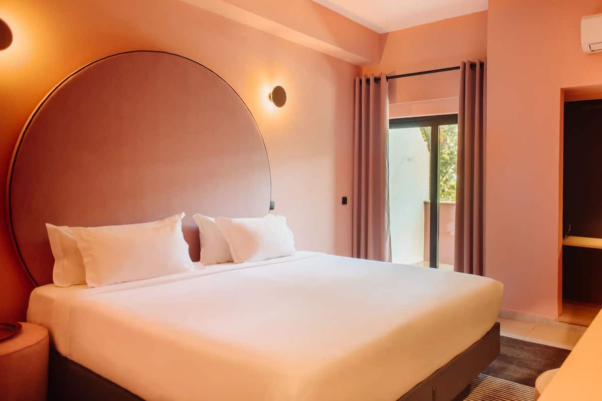Een hotelkamer met een groot bed, een rond hoofdeinde, een nachtkastje, een raam met gordijnen en een deur.