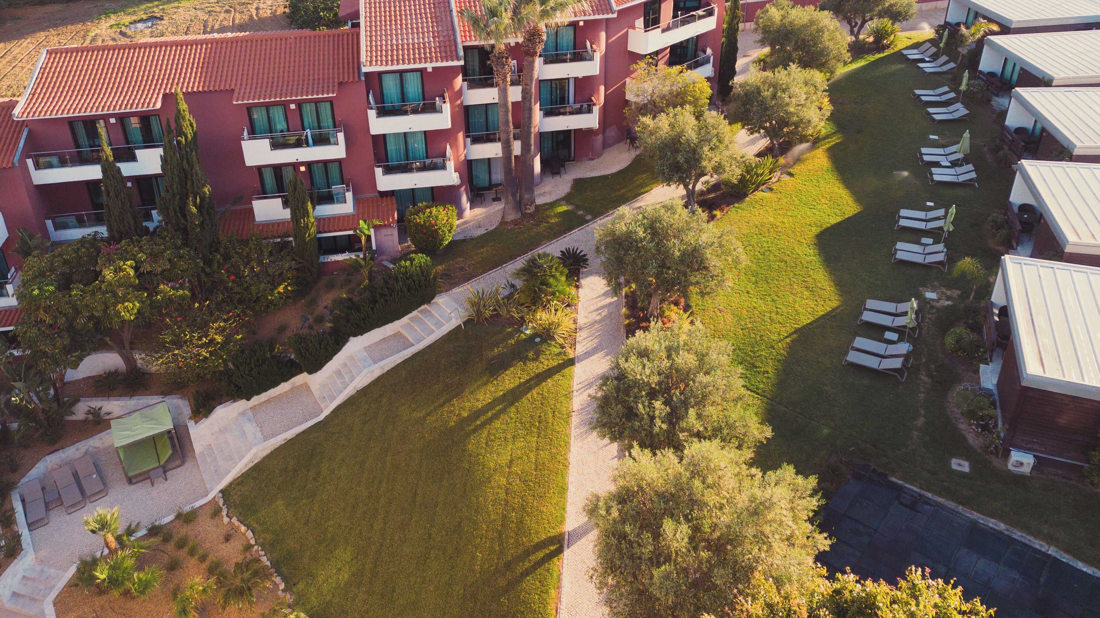 A multi-story hotel with balconies, a central courtyard with grass and trees, and a pool area with sun loungers.