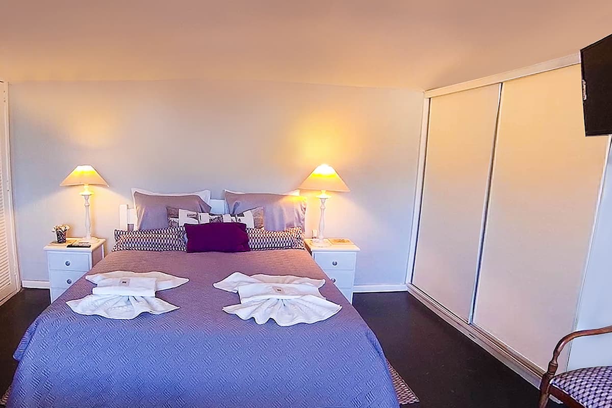 A hotel room with a large bed, two bedside lamps, a wardrobe, a chair, and a nightstand with a lamp.