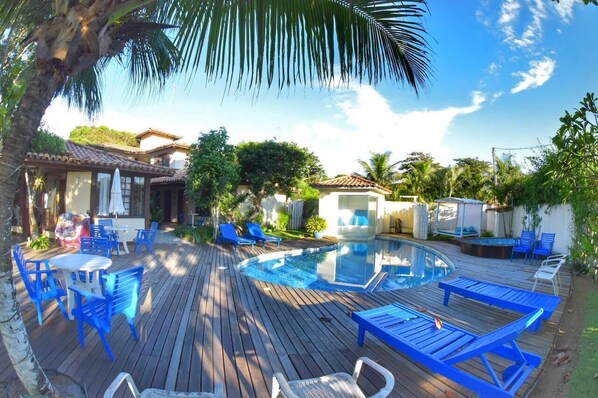 Terraza de madera con piscina, sillas reclinables azules y un área de estar cubierta.