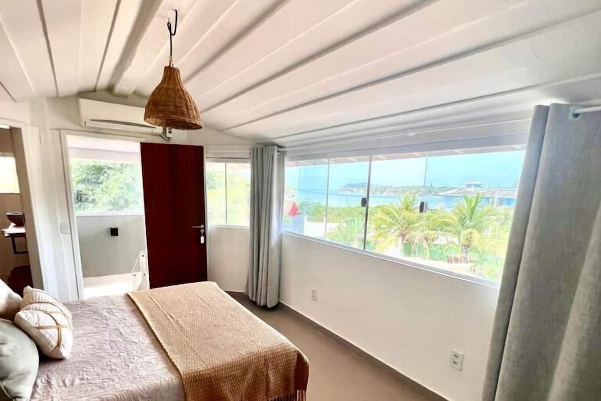 A bedroom with a bed, a hanging light fixture, a window with a view, and a door leading to another room.