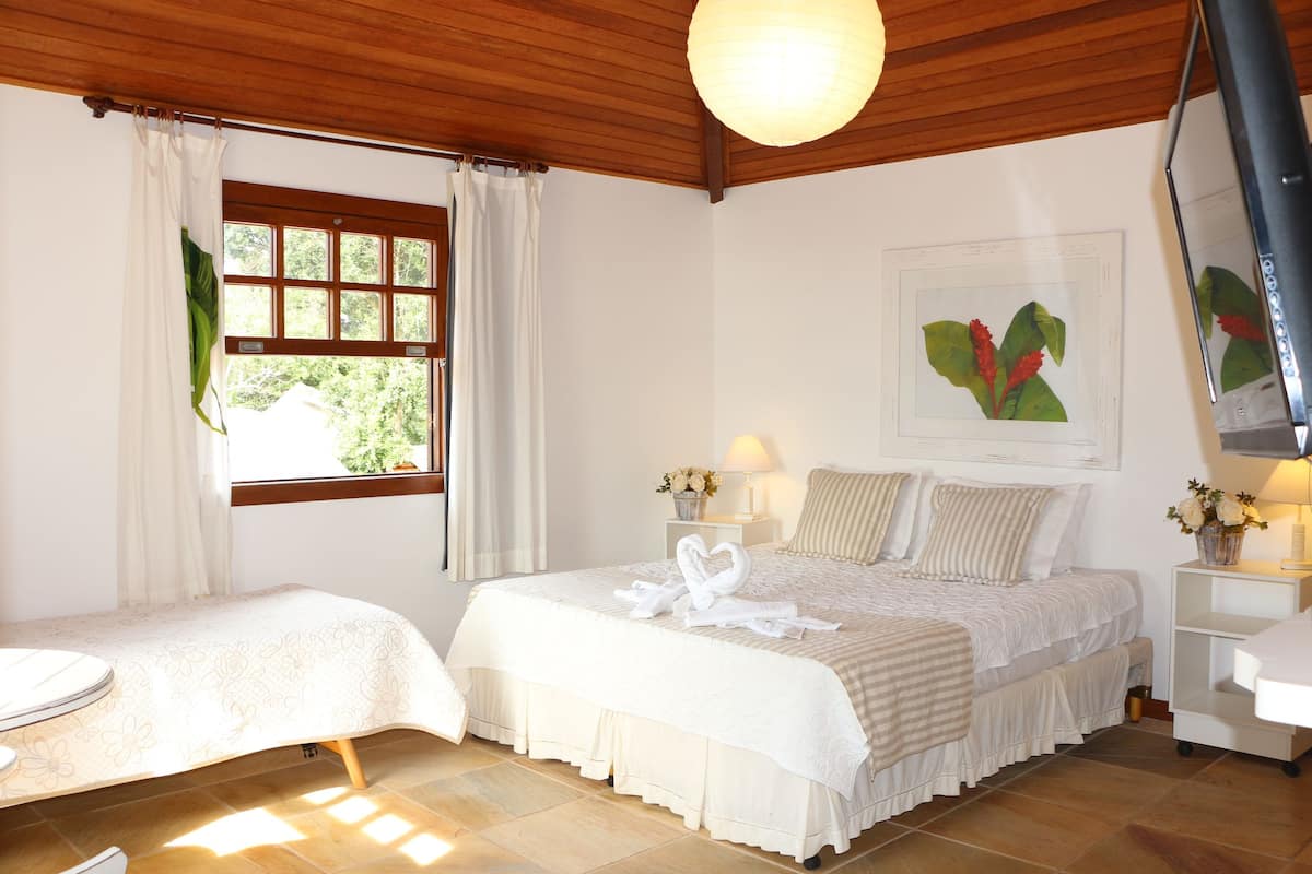 A hotel room with two beds, a large window with curtains, a wooden ceiling, a wall-mounted lamp, and a framed botanical print.